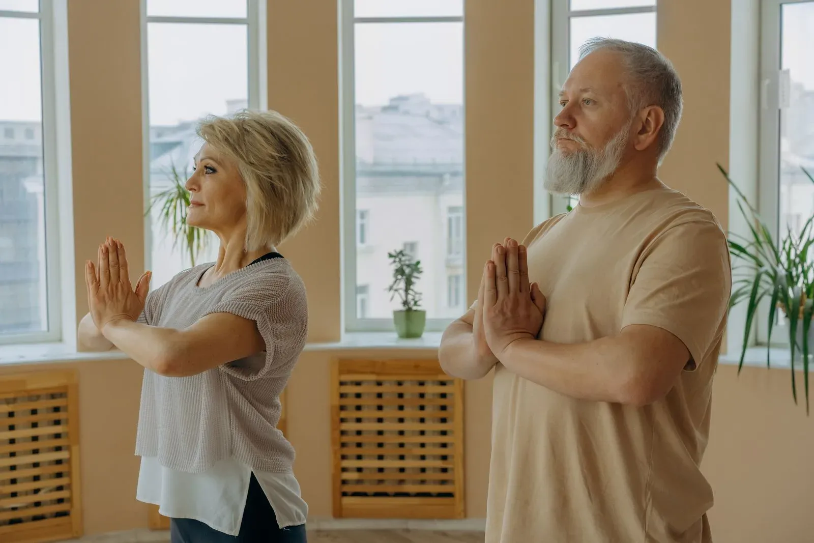 Casal 60+ praticando yoga em pé, em posição de oração, num ambiente iluminado em casa Casal 60+ praticando yoga em pé, em posição de oração, num ambiente iluminado em casa