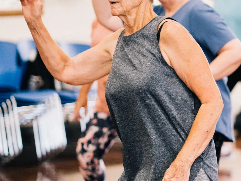 Mulher 60+ dançando com o braço erguido em aula de fitness em salão comunitário
