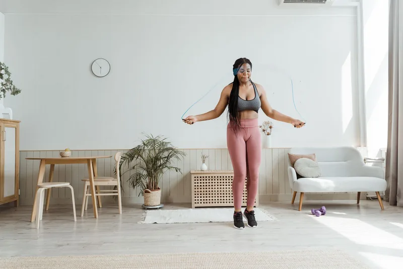 Mulher pulando corda no meio da sala de casa, halteres apoiados no chão ao fundo Mulher pulando corda no meio da sala de casa, halteres apoiados no chão ao fundo