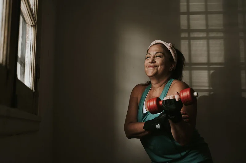 Mulher 40+ sorrindo durante rosca bíceps com halteres em casa Mulher 40+ sorrindo durante rosca bíceps com halteres em casa