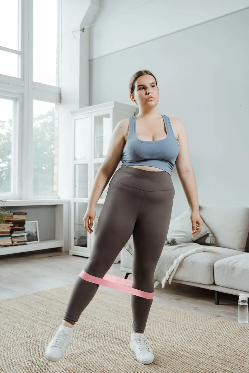 Mulher em pé na sala de casa com elástico de resistência posicionado nas pernas para exercício de panturrilha Mulher em pé na sala de casa com elástico de resistência posicionado nas pernas para exercício de panturrilha