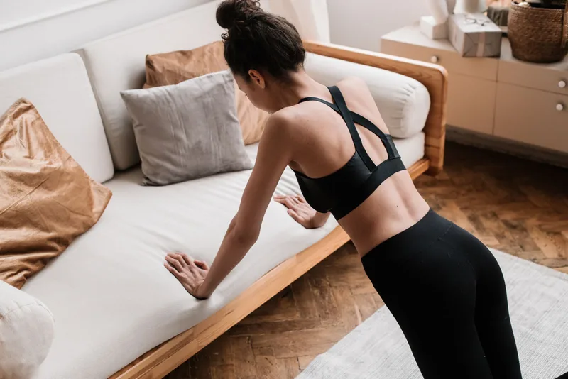 Mulher apoiando as mãos no sofá para fazer flexão inclinada, ativando o ombro com o próprio peso corporal em casa Mulher apoiando as mãos no sofá para fazer flexão inclinada, ativando o ombro com o próprio peso corporal em casa