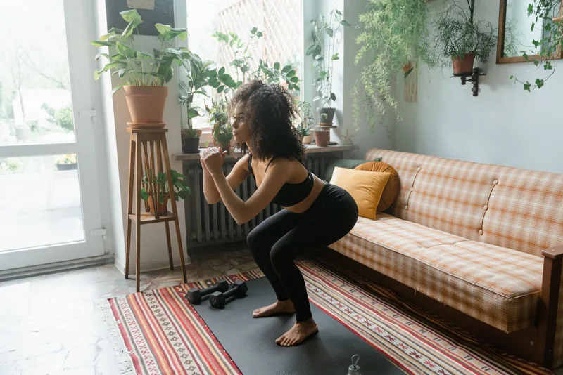 Mulher fazendo agachamento na sala de casa, halteres apoiados no tapete ao lado Mulher fazendo agachamento na sala de casa, halteres apoiados no tapete ao lado