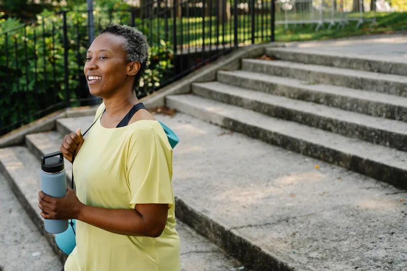 Mulher 60+ sorrindo, com tapete de exercícios e garrafa de água, a caminho do treino Mulher 60+ sorrindo, com tapete de exercícios e garrafa de água, a caminho do treino