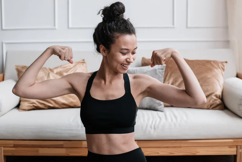 Mulher sorrindo e flexionando os braços na sala de casa, após o treino Mulher sorrindo e flexionando os braços na sala de casa, após o treino