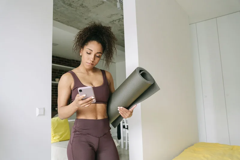 Mulher conferindo o cronograma de treino no celular, tapete de exercícios em mãos Mulher conferindo o cronograma de treino no celular, tapete de exercícios em mãos