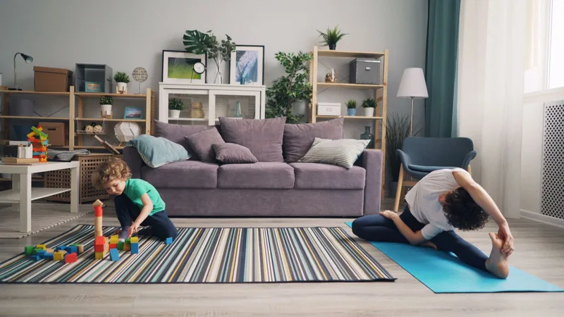 Mãe alongando no tapete de exercícios enquanto o filho brinca com blocos no mesmo cômodo