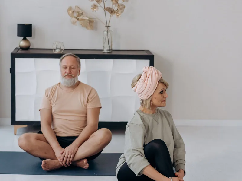 Casal 60+ praticando yoga sentado no chão, um em postura de meditação e outro em torção sentada Casal 60+ praticando yoga sentado no chão, um em postura de meditação e outro em torção sentada