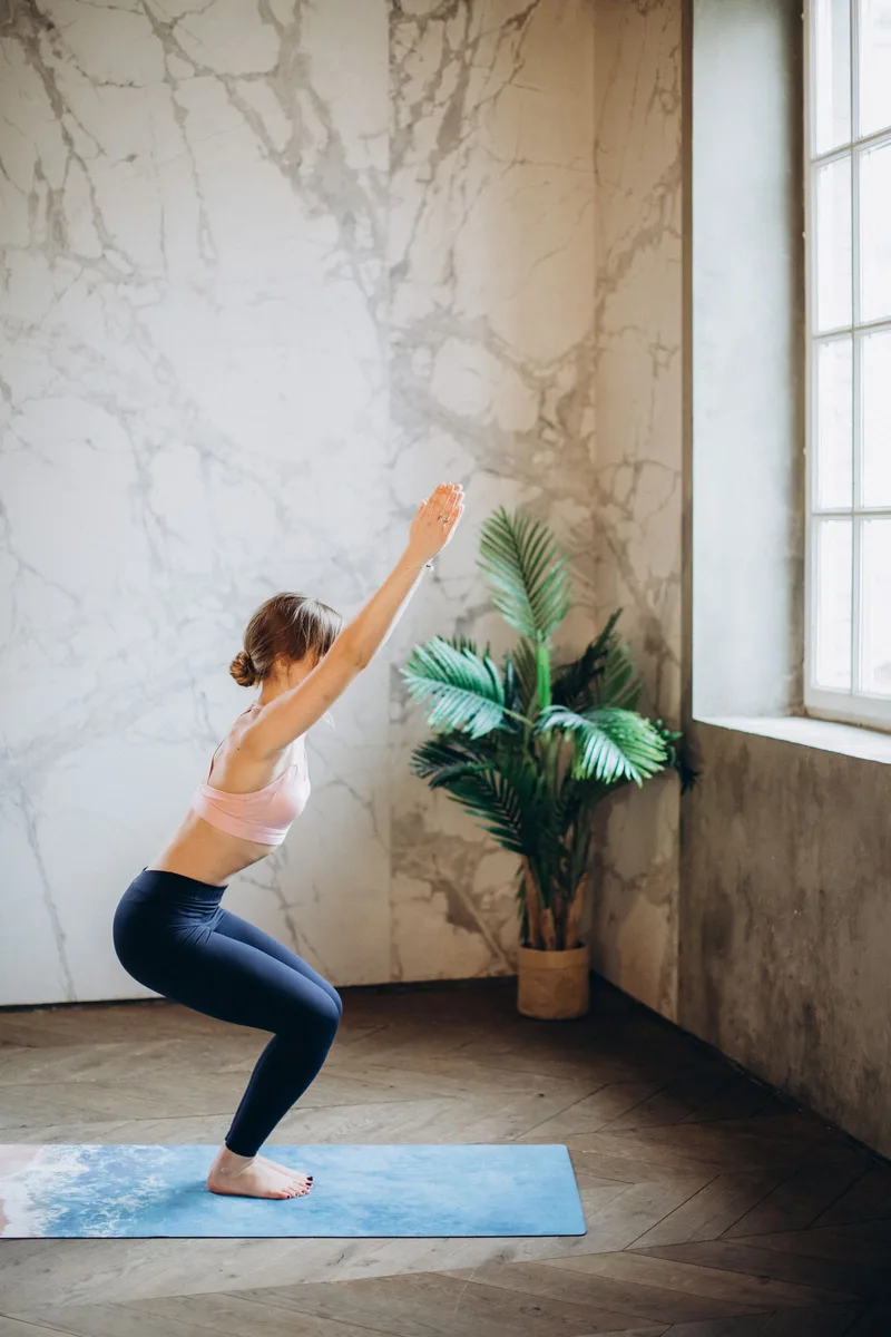Mulher em Utkatasana, joelhos flexionados como se fosse sentar em uma cadeira, braços erguidos